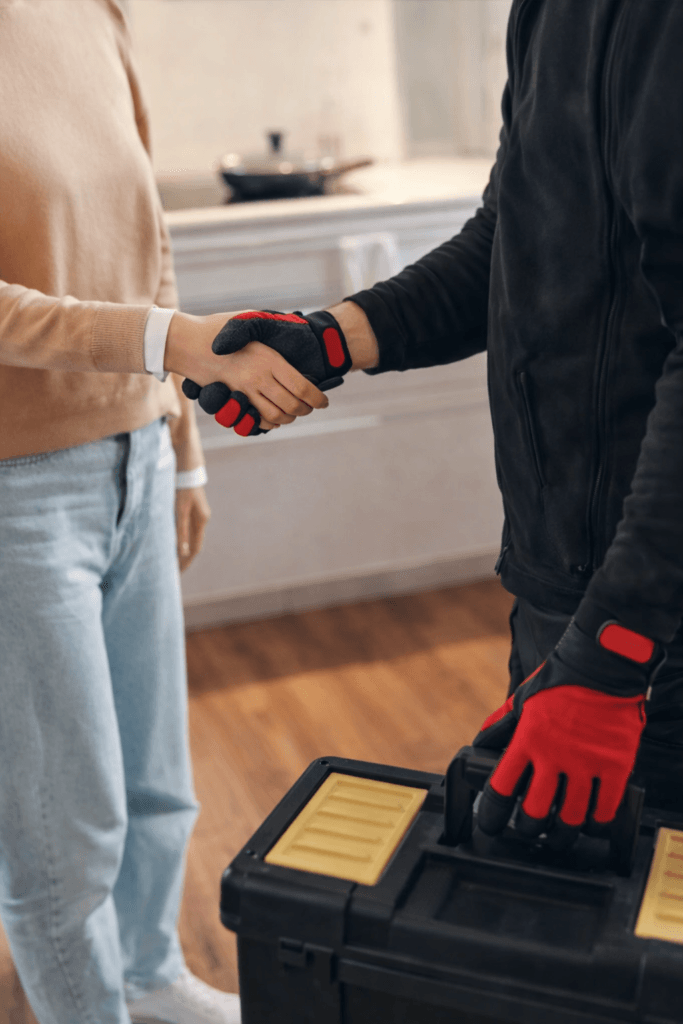 Close-up of a homeowner shaking hands with a tradesperson wearing red-and-black work gloves, who is holding a black toolbox in a bright kitchen.